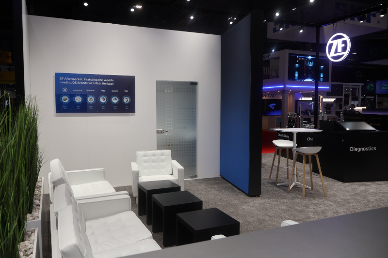 A semi-private lounge area inside the ZF booth with contemporary furniture, wood-textured flooring, and a glass-walled meeting room for professional B2B consultations.